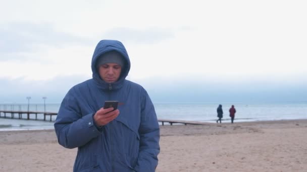 Hombre en una chaqueta en la playa junto al mar en invierno hablando por teléfono móvil . — Vídeos de Stock