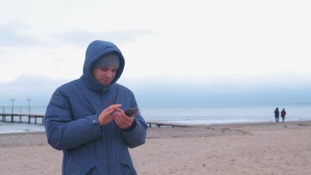 Man in a blue down jacket on the sand beach by the sea typing a message on mobile phone. — Stock Video