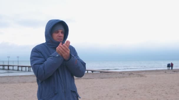 Mann in Jacke am Meer wärmt gefrorene Hände und wartet auf jemanden. — Stockvideo