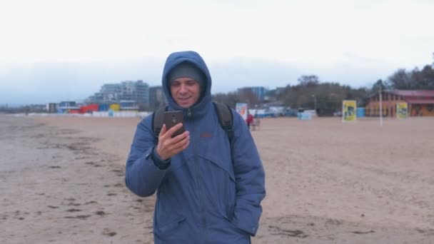 Homem blogueiro em uma jaqueta azul para baixo andando na praia de areia junto ao mar e falando um bate-papo por vídeo no celular . — Vídeo de Stock