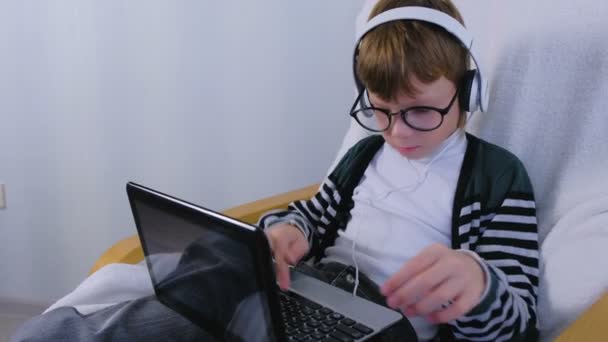 Boy writes and types a message on laptop in headphones and glasses sitting in armchair. — Stock Video