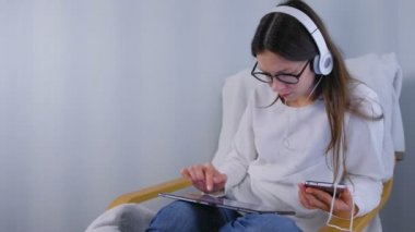 Kadın gözlük ve kulaklık smartphone ve koltukta oturan tablet kullanarak çevrimiçi mağazada alışveriş yapar.