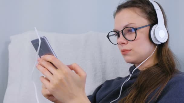 メガネの女性がスマート フォンを自宅でヘッドフォンで音楽をリッスンします。. — ストック動画