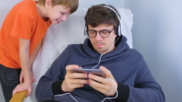 Homme en lunettes et écouteurs jouant à un jeu sur smartphone avec son fils. Gamer relax . — Video