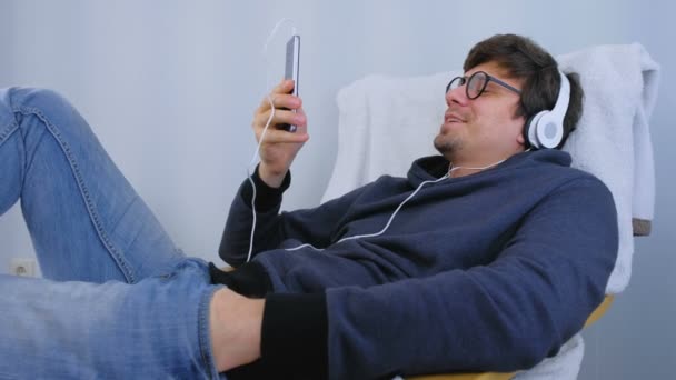 El hombre escucha música en los auriculares en el teléfono inteligente y canta una canción en el karaoke. Tiempo de relax en sillón . — Vídeos de Stock