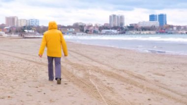 Parlak sarı ceketli adamı deniz kıyısında kumlu bir plaj üzerinde arkadan görünüm yürür..