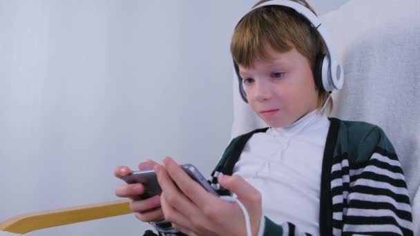 Boy está jugando un juego en los auriculares en el teléfono inteligente sentado en el sillón en casa . — Vídeos de Stock