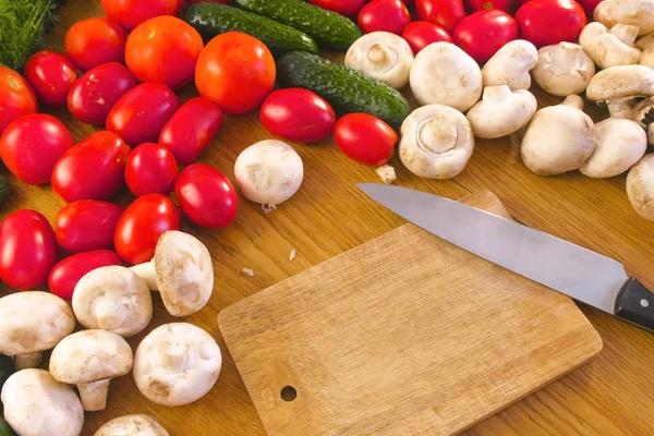 Tablero de madera con cuchillo y verduras frescas tomates, pepinos, calabacines, pimientos, verduras, rábanos y champiñones en la mesa de la cocina . — Foto de Stock