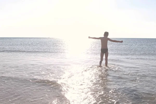 Chłopiec sylwetka jest chodzenie przez wody morza na zachód słońca. Pływanie w morzu. — Zdjęcie stockowe