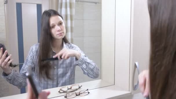 Mujer despertada se peina el pelo de pie frente a un espejo en el baño y mirando el teléfono móvil . — Vídeos de Stock