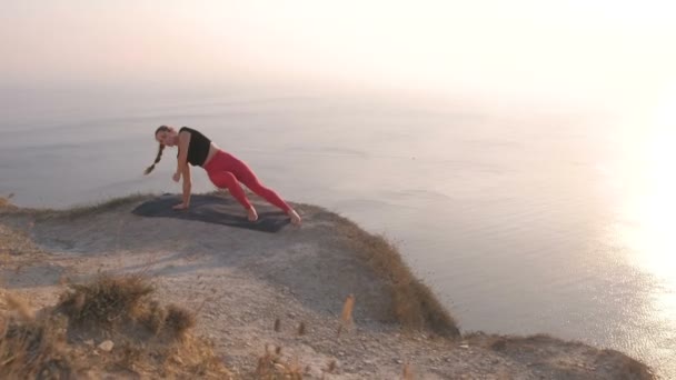 Hermosa vista de la mujer haciendo yoga nantasana, elevación lateral de la pierna en la montaña con vista al mar al atardecer . — Vídeo de stock