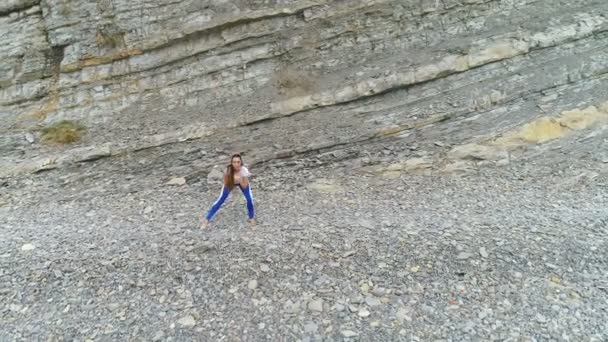 Kvinnan gör knäböj bodyflex under andningsövningar på havet och rock bakgrunden. Aerial vackra bilder Visa. — Stockvideo