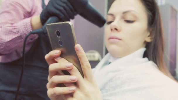 Holič sušení vlasů s vysoušečem vlasů. Posiluje vlasy s keratinem. Klient se dívá na mobilní telefon. — Stock video