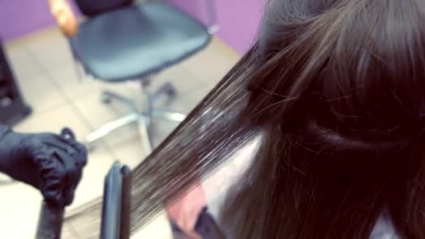 Coiffeur redresse les cheveux brun foncé de belle femme en utilisant des pinces à cheveux dans le salon de beauté. Gros plan sur les cheveux . — Video