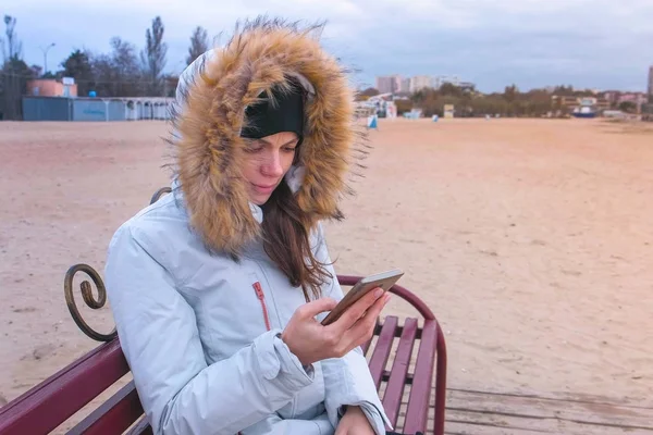Женщина Белой Куртке Сидит Скамейке Пляже Смотрит Грустное Видео Мобильный — стоковое фото