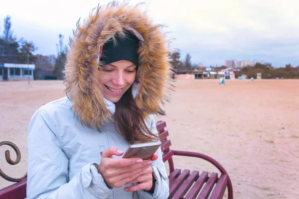 Mujer en una chaqueta blanca sentada en un banco en la playa y mirando el vídeo increíble en el teléfono móvil . — Foto de Stock