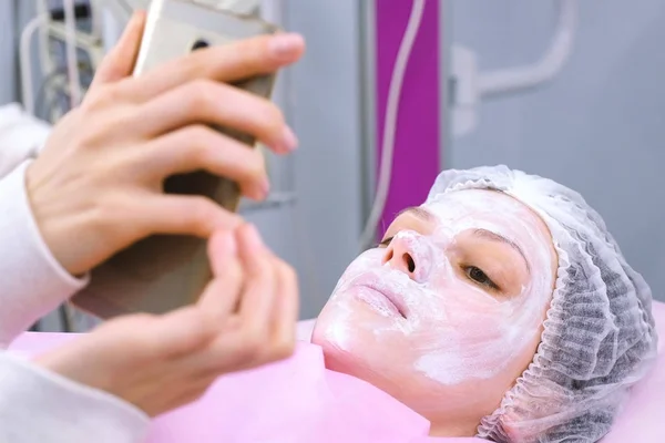 Donna legge qualcosa in un telefono cellulare sdraiato sulla procedura nell'ufficio estetiste con una maschera sul viso . — Foto Stock