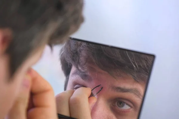 Young man plucks his eyebrows with tweezers in front of the mirror at home.