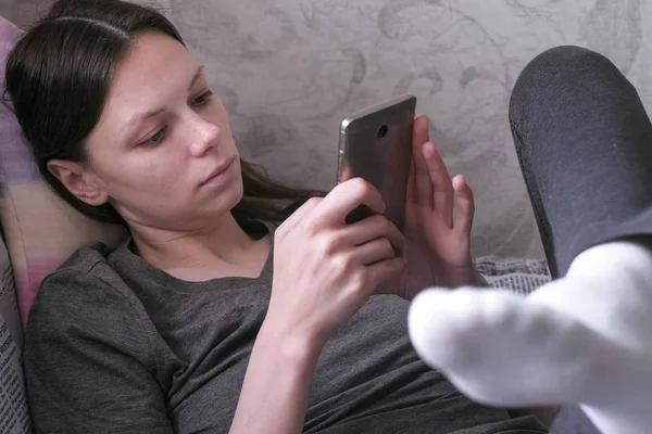 Сонная женщина лежит на диване и читает что-то по мобильному телефону . — стоковое фото