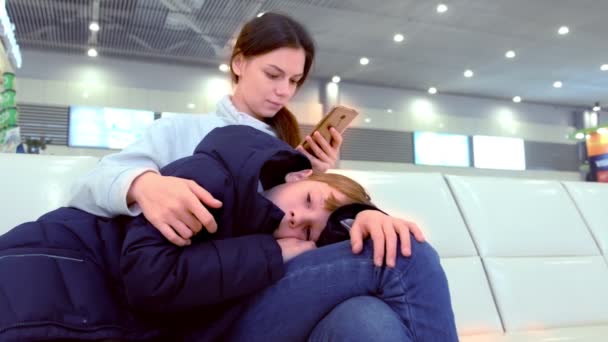 Femme avec son fils fatigué enregistrement en ligne sur son téléphone portable dans le hall de l'aéroport . — Video