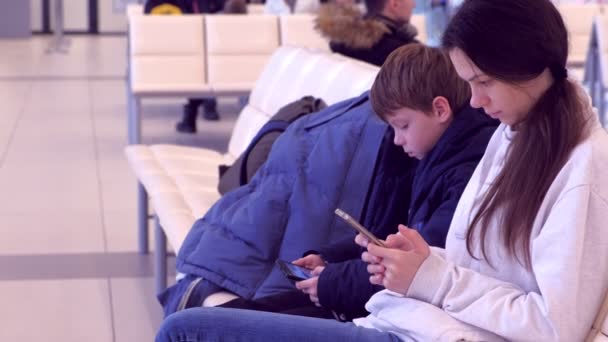 Женщина с сыном просматривает мобильные телефоны в зале ожидания рейса, вид сбоку . — стоковое видео