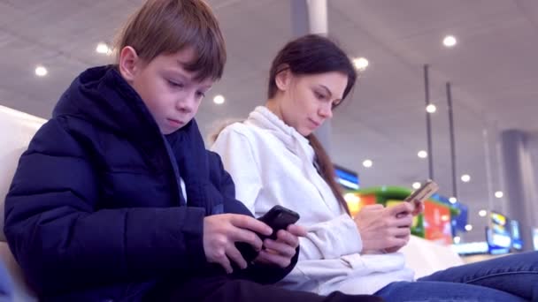 Mulher com seu filho jogando jogos em telefones celulares no corredor do aeroporto à espera de voo . — Vídeo de Stock