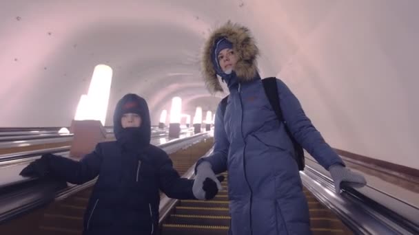 Mamá e hijo en ropa de invierno se están moviendo por la escalera mecánica en el túnel del metro . — Vídeos de Stock