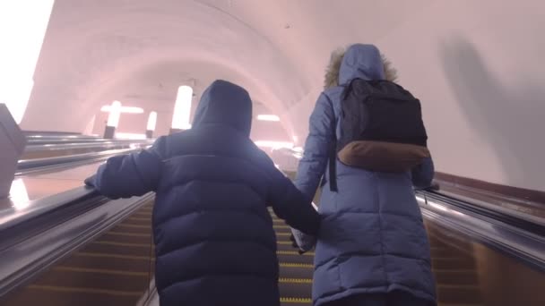 Mamá y su hijo en ropa de invierno se están moviendo por la escalera mecánica en el túnel del metro, vista trasera . — Vídeos de Stock