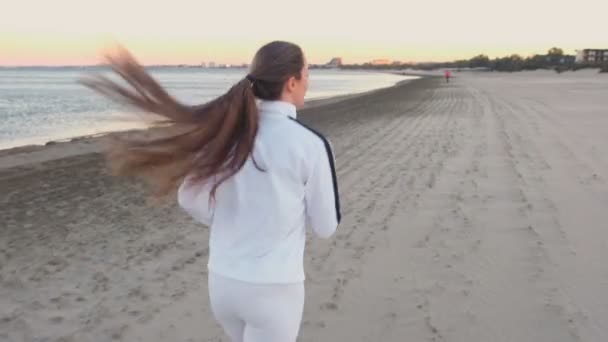 Junge Frau joggt bei Sonnenaufgang im Herbst am Sandstrand am Meer, Rückansicht. — Stockvideo