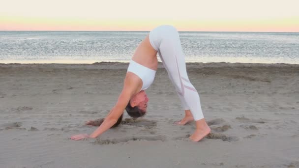 Jovem está fazendo ioga Adho mukha shvanasana na praia de areia ao nascer do sol . — Vídeo de Stock