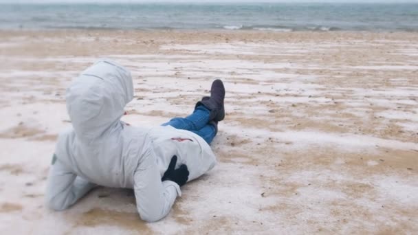 Kvinna turist ligger på en snöig sandstrand på vintern vid havet. Kvinna i en jacka och jeans, bakifrån. — Stockvideo