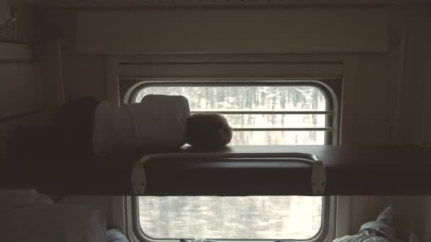Pojke på tåget liggande på översta hyllan i en andra klassens vagn och tittar på fönster. Bakifrån. — Stockvideo
