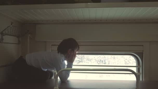 Niño en el tren acostado en el estante superior en un carro de segunda clase y mirando a la ventana. Vista lateral . — Vídeos de Stock