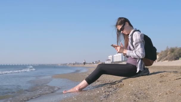 Chica viajera con una mochila sentada en una playa de arena y escribiendo un mensaje en el teléfono móvil . — Vídeos de Stock