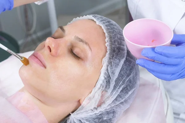 La esteticista pone una máscara en la cara de las mujeres con un cepillo. Manos de un cosmetólogo con guantes de goma azul. Tratamientos faciales. Vista lateral . — Foto de Stock