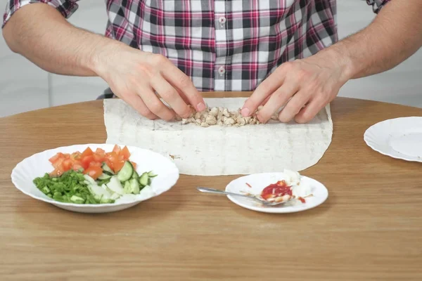 Мужчина готовит шаурму на кухонном столе дома. Пита, овощи и зеленый лук с соусом и майонезом. Руки крупным планом . — стоковое фото