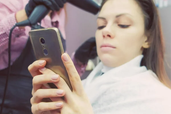 Barbeiro secando o cabelo com secador de cabelo. Fortalecendo o cabelo com queratina. Cliente olha para o telefone móvel . — Fotografia de Stock