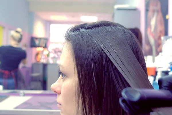 Peluquería alisados cabello castaño oscuro de mujer hermosa usando pinzas para el cabello en el salón de belleza. Vista lateral . — Foto de Stock