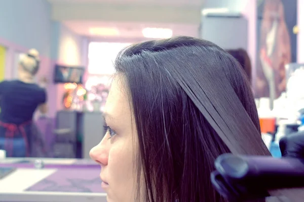 Hairdresser straights dark brown hair of beautiful woman using hair tongs in beauty salon. Side view. — Stock Photo, Image