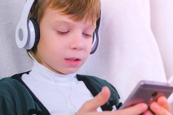 Boy está escuchando música en los auriculares en el teléfono inteligente y cantando una canción sentada en el sillón en casa . — Foto de Stock