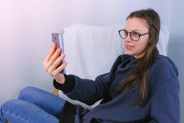 Счастливая юная брюнетка в очках разговаривает в видеочате по мобильному телефону, сидя в кресле . — стоковое фото