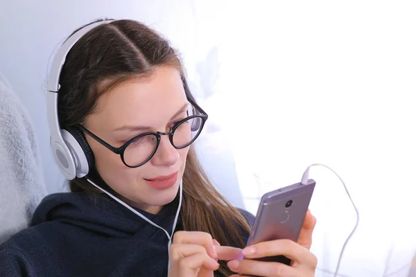 Happy young brunette woman in glasses and headphones talk a video chat on a mobile phone. — Stock Photo, Image