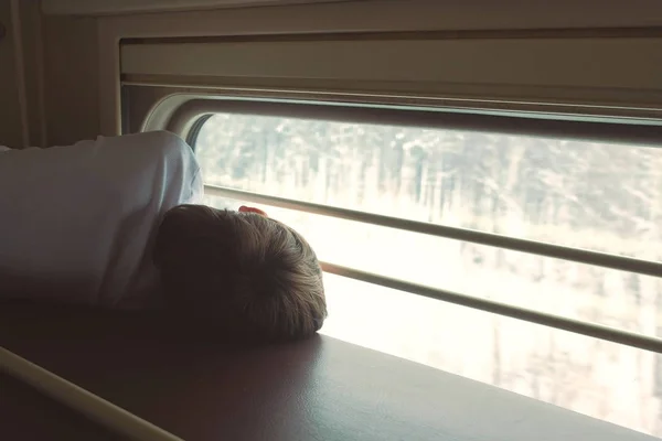 Rapaz no comboio deitado na prateleira de cima numa carruagem de segunda classe e a olhar para a janela. Visão traseira . — Fotografia de Stock