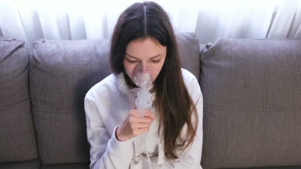 Använda nebulisatorn och inhalator för behandling. Unga kvinnan andas in genom inhalatorn masken sitter på soffan. — Stockvideo
