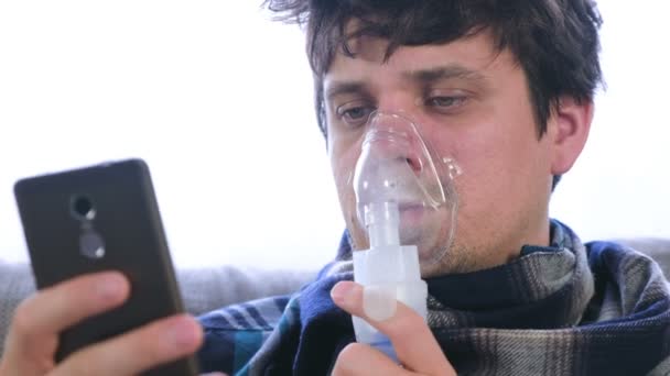 Use nebulizador e inhalador para el tratamiento. Hombre enfermo inhalando a través de la máscara del inhalador y mirando el teléfono móvil sentado en el sofá . — Vídeos de Stock