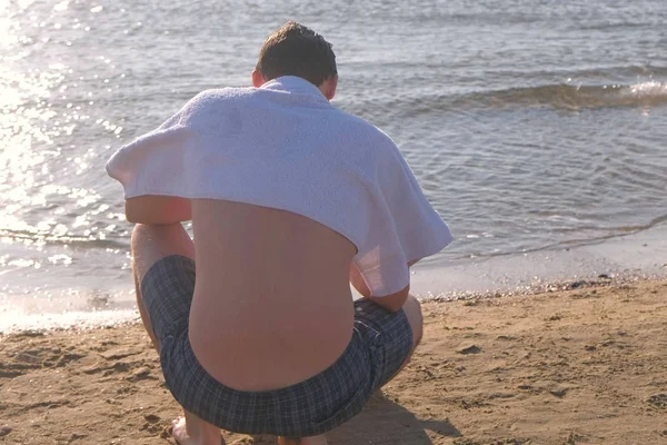 El hombre está sentado en la playa, cubriéndose los hombros con una toalla del sol . — Foto de Stock