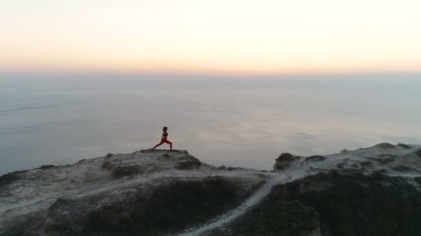 在日落时 女人正在山顶上做瑜伽 可以看到海景 空中镜头 侧视图 — 图库视频影像