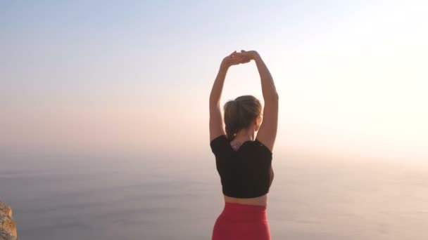 Hermosa vista de mujer haciendo yoga estirándose en la montaña con vista al mar al atardecer. Levantando las manos. Vista trasera . — Vídeo de stock