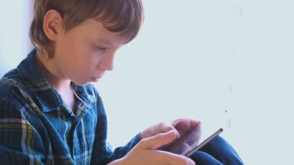 Boy está jugando juegos en la tableta sentado en el alféizar de la ventana . — Vídeos de Stock