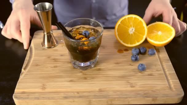 Cóctel de alcohol en el vaso de rocas con naranja, hielo y arándanos en el tablero de madera, manos de cerca . — Vídeo de stock